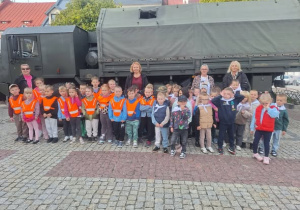 dzieci i nauczycielki w kamizelkach odblaskowych stoją na tle wojskowego samochodu ciężarowego