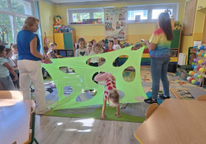 Dzieci przechodzą przez materiał z dziurami. Dwie kobiety trzymają materiał za ranty
