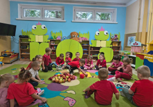 dzieci siedzące w kole ubrane na czerwono. Na środku koła jabłka i chłopiec trzymający kostkę do gry