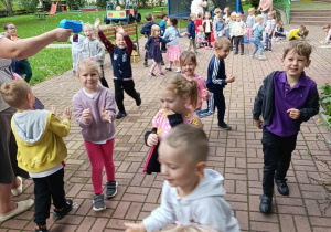Grupa dzieci bawiąca się w ogrodzie przedszkolnym. Część dzieci biega inne uśmiechnięte podskakują.