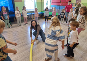 Grupa dzieci stojąca w kole, nauczycielka trzymająca w dłoni makaron do pływania. Dzieci przeskakują przez makaron.