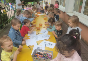 Grupa dzieci siedząca przy stoliku wykonująca bransoletki, nawlekając koraliki na sznurek.