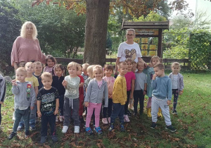 Grupa dzieci z dwiema Paniami, stojąca w ogrodzie przedszkolnym.