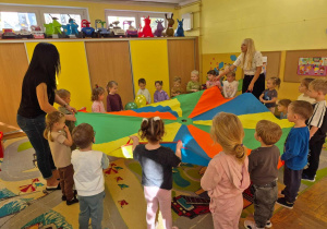 Grupa dzieci wraz z Paniami trzymająca kolorową chustę animacyjną w sali przedszkolnej.
