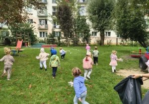 Grupa dzieci zbierająca śmieci w ogrodzie przedszkolnym. Pani trzymająca czarny worek na śmieci.
