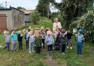 Grupa dzieci wraz z Panią nauczycielką. Trzymająca dłonie w górze pokazując foliowe rękawiczki do zbierania śmieci.