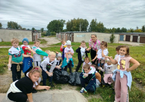 Grupa dzieci wraz z nauczycielką, pokazująca palcem na worki pełne śmieci, robiąca śmieszne miny.