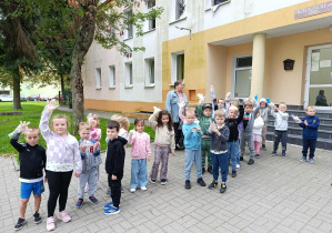 Grupa dzieci stojąca przed przedszkolem. Dzieci trzymają w górze dłonie. Niektóre pokazują rękawiczki foliowe do zbierania śmieci.