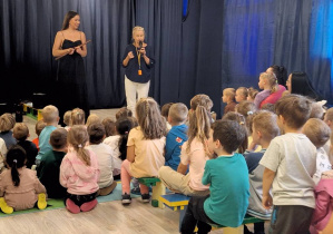 Dzieci siedzące tyłem patrzą na kobietę trzymającą flet oraz kobietę trzymającą mikrofon