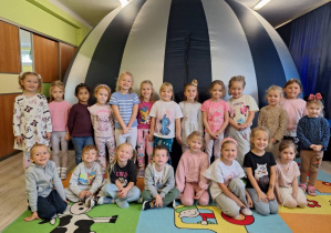 Grupa dzieci z nauczycielką siedząca i stojąca na tle kopuły planetarium.
