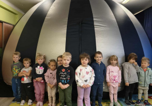 Grupa dzieci z nauczycielką siedząca i stojąca na tle kopuły planetarium.