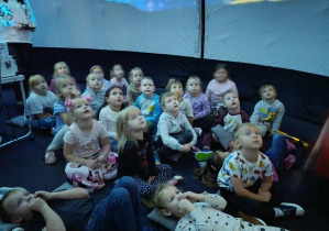 Grupa dzieci siedząca w kopule planetarium, oglądająca wyświetlający się film.