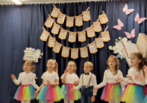 Dziewczynki w białych bluzkach i kolorowych spódniczkach i chłopiec ubrany na galowo ilustrują słowa piosenki.