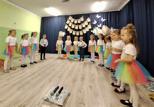 Dziewczynki w białych bluzkach i kolorowych spódniczkach i chłopcy ubrany na galowo ilustrują słowa piosenki.