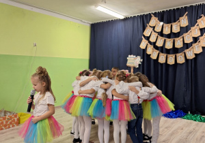 Grupa dzieci obejmująca się w ramionach i tworząca koło. Przed nimi stoi dziewczynka z mikrofonem.