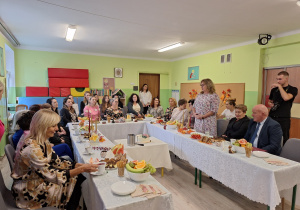 Grupa dorosłych osób siedząca przy stole na którym znajdują się dekoracje jesienne oraz poczęstunek. Kobieta stoi i przemawia do grupy ludzi.