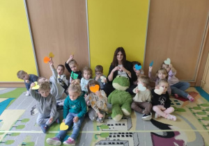 Grupa dzieci siedząca na dywanie z panią nauczycielką. Każde trzyma kolorowe serce, niektóre dzieci unoszą je w górę.