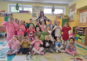 Grupa dzieci pokazująca serca z dłoni. Część z nich siedzi na dywanie, część stoi. Za nimi jest nauczycielka pokazująca znak serca z dłoni.