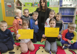 Grupa dzieci siedząca na dywanie z nauczycielką, dwie dziewczynki trzymają kartkę z napisem "Dzień życzliwości". Pani nauczycielka obejmuje chłopców do zdjęcia.