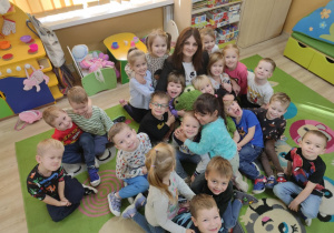 Grupa dzieci wraz z nauczycielką siedząca na dywanie przytulająca się do maskotki zielonej żaby, patrząca do zdjęcia.