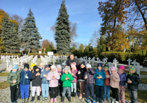 Grupa dzieci z grupy "Misie" stojąca z nauczycielką, trzymająca w rękach znicze, na tle grobów poległych żołnierzy.
