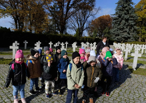 Grupa dzieci z grupy "Pszczoły", stojąca na tle grobów poległych żołnierzy, uśmiechająca się do zdjęcia.