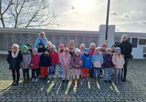 Grupa dzieci z grupy "Słoneczka", stojąca wraz z nauczycielkami, uśmiechająca się do zdjęcia, na tle tablicy z imionami poległych żołnierzy.
