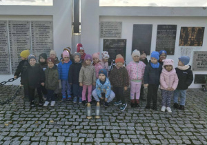 Grupa dzieci z grupy "Żaby", stojąca na tle tablicy z imionami poległych żołnierzy. Przed nimi stoją dwa białe znicze.