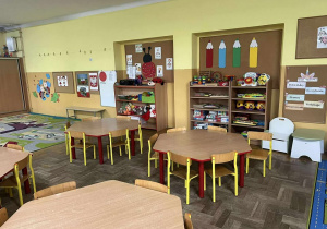 Na pierwszym planie widać stoliki z krzesłami, na drugim półki z zabawkami oraz kolorowe tablice