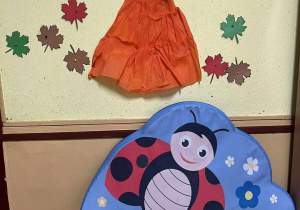 Na pierwszym planie półki na zabawki, materac z wizerunkiem biedronki, na drugim planie dekoracja, wycięta postać pani jesieni z kolorowymi liśćmi