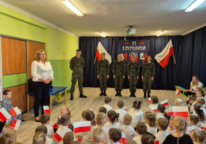Pani Dyrektor Przedszkola, przemawiająca do zgromadzonych dzieci, dzieci siedzące na dywanie, ubrane na galowo, trzymające flagi Polski, na drugim planie młodzież w mundurach