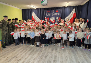 Zdjęcie grupowe dzieci ubranych na galowo, trzymających flagi, po prawej stronie stojące opiekunki, po lewej stronie młodzież w mundurach, na tle dekoracji z okazji 11 listopada