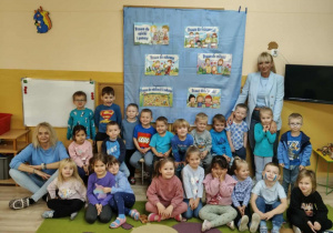 Grupa dzieci ubrana na niebiesko. Cześć dzieci siedzi na dywanie pozostałe dzieci siedzą na ławce. Obok stoją nauczycielki. W tle niebieski materiał ze zdjęciami.