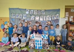 Grupa dzieci ubrana na niebiesko. Cześć dzieci siedzi na dywanie pozostałe dzieci siedzą na ławce. Obok stoją nauczycielki. Dzieci trzymają w dłoniach prace plastyczną dużego formatu. .