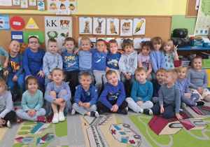 Grupa dzieci ubrana na niebiesko. Cześć dzieci siedzi na dywanie pozostałe dzieci siedzą na ławce. Dzieci uśmiechają się.