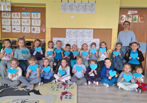 Grupa dzieci ubrana na niebiesko. Cześć dzieci siedzi na dywanie pozostałe dzieci siedzą na ławce. Obok stoją nauczycielki. Dzieci trzymają w dłoniach niebieskie serca.