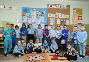 rupa dzieci ubrana na niebiesko. Cześć dzieci siedzi na dywanie pozostałe dzieci siedzą na ławce. Dzieci uśmiechają się.