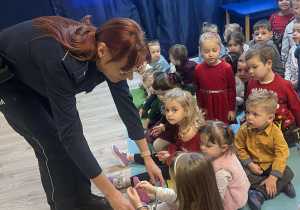 Pani policjantka zakłada dziecku kajdanki na dłonie. Grupa dzieci przygląda się policjantce.