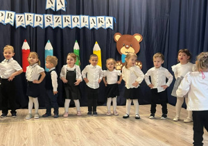 Grupa dzieci ubrana na galowo trzymającą się za biodra. Dzieci tańczą i śpiewają.