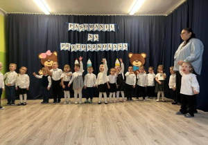 Grupa dzieci ubrana na galowo trzymającą dłonie w górze. Dzieci tańczą i śpiewają.