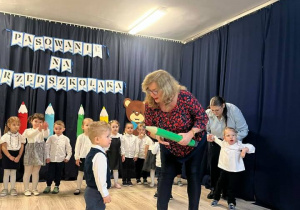 W tle stoją dzieci ubrane na galowo. Z przodu stoi chłopiec, kobieta dotyka jego ramienia dużą kredką.