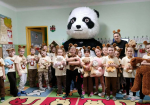 Dzieci ubrane na brązowo z uszami misiów stoją obok wielkiego misa pandy. Dzieci uśmiechają się.