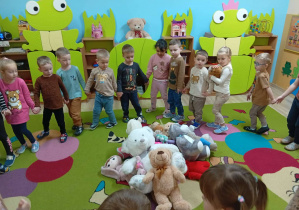 Dzieci stoją w kole na środku leżą pluszowe misie.