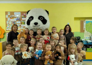 Dzieci ubrane na brązowo stoją obok wielkiego misa pandy. Dzieci uśmiechają się.