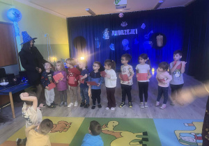Dzieci stoją z kartkami z wróżami w dłoniach wiedźma stoi obok.