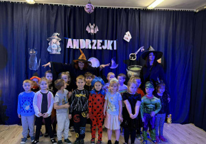 Dzieci stoją na tle napisu ,,Andrzejki'' obok stoi wiedźma.