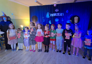 Dzieci stoją z kartkami z wróżami w dłoniach wiedźma stoi obok.