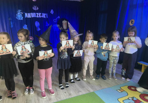 Dzieci stoją z kartkami z wróżami w dłoniach wiedźma stoi obok.