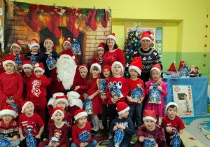 Grupa dzieci w czerwonych strojach i czapkach Mikołajów. Dzieci trzymają w dłoniach torebki prezentowe.