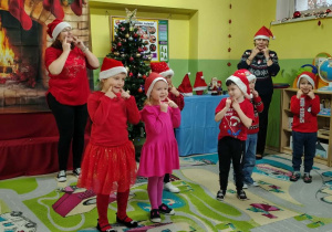 Grupa dzieci w czerwonych strojach i czapkach Mikołajów. Dzieci tańczą i pokazują za twarze.